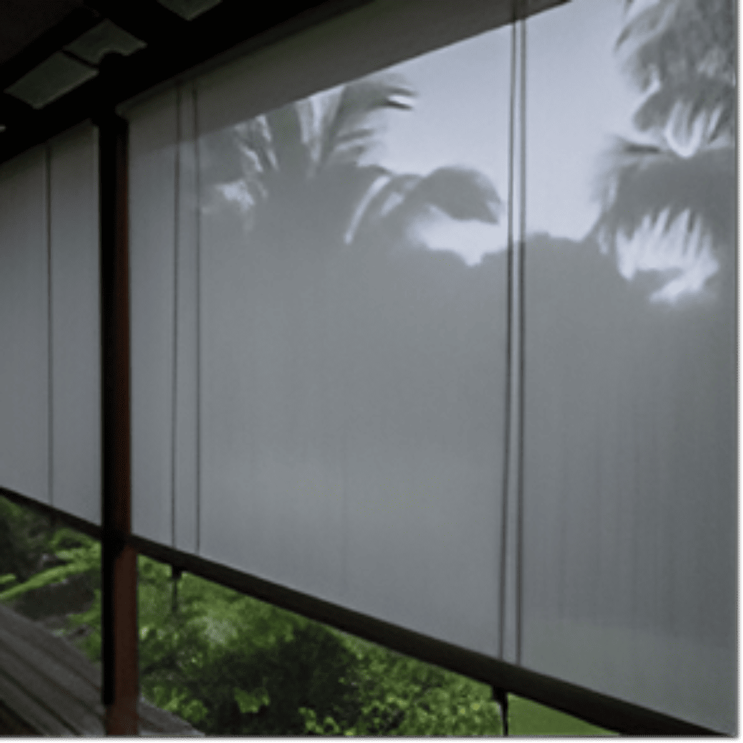 A set of fabric monsoon blinds, a durable and translucent grey material, is rolled down on a veranda during a downpour. Rainwater streaks down the surface of the blinds, and a blurred, lush tropical garden is visible through the material. 