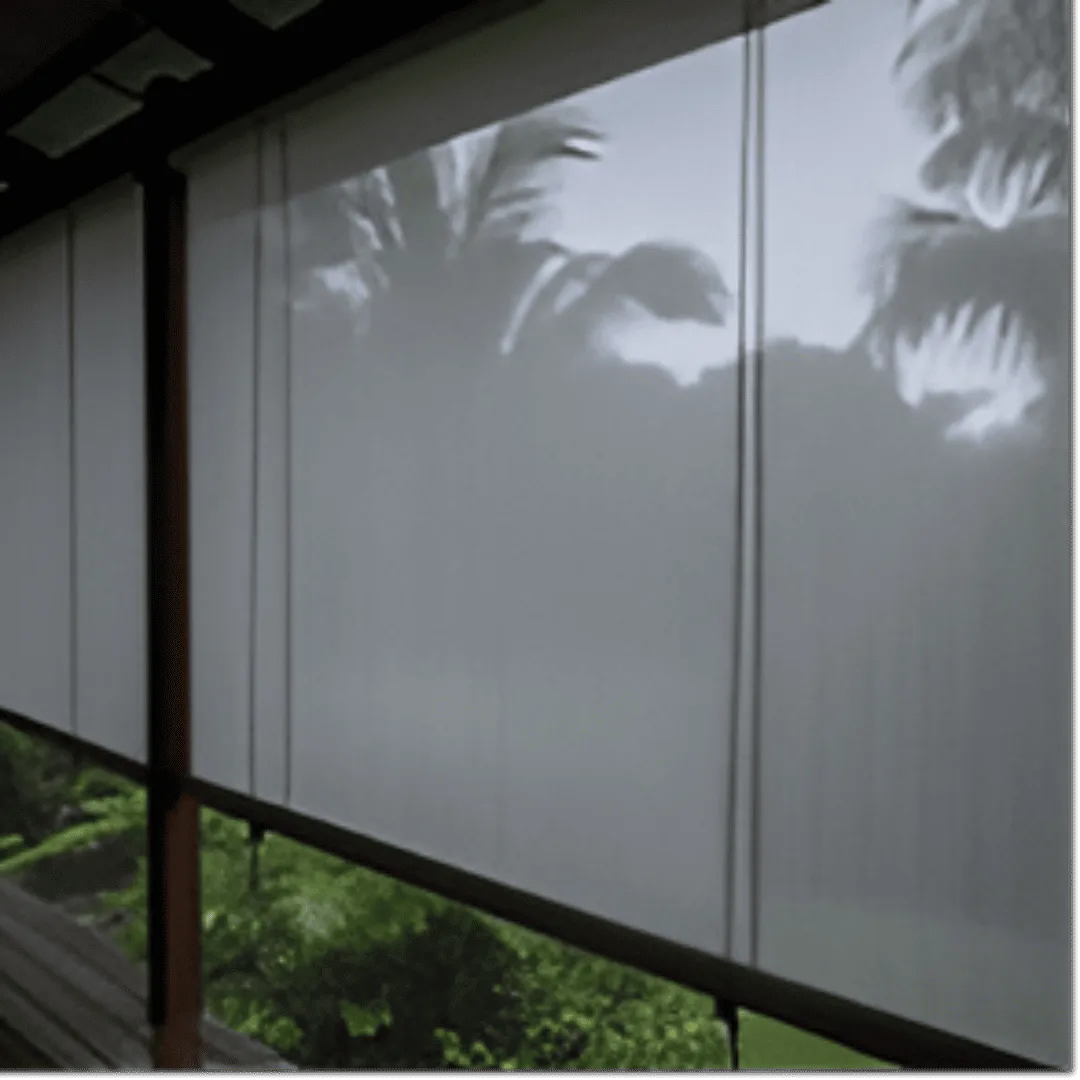 A set of fabric monsoon blinds, a durable and translucent grey material, is rolled down on a veranda during a downpour. Rainwater streaks down the surface of the blinds, and a blurred, lush tropical garden is visible through the material. 