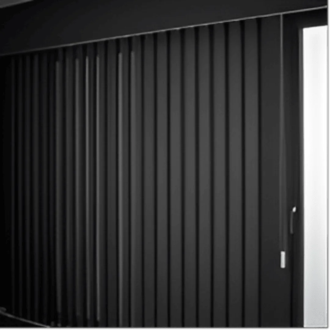 A close-up view of modern, sleek blackout vertical blinds installed on a large window. The blinds are made of a thick, opaque fabric in a dark charcoal grey, effectively blocking out all light. They are perfectly aligned and slightly overlapping. .