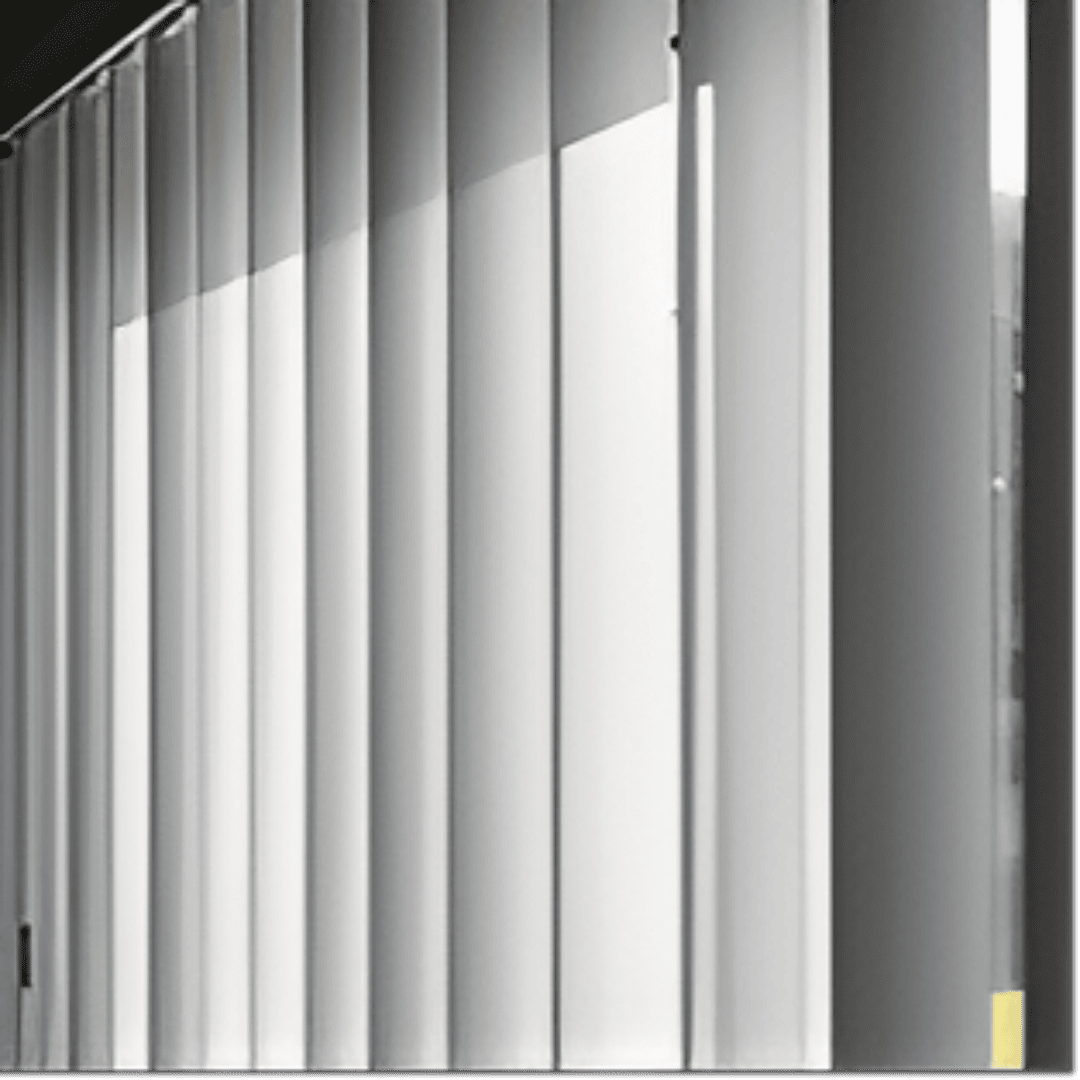 A close-up, eye-level shot of white PVC vertical blinds, partially open, with sunlight streaming through them from the left side. The individual slats are slightly angled, creating alternating strips of light and shadow. 