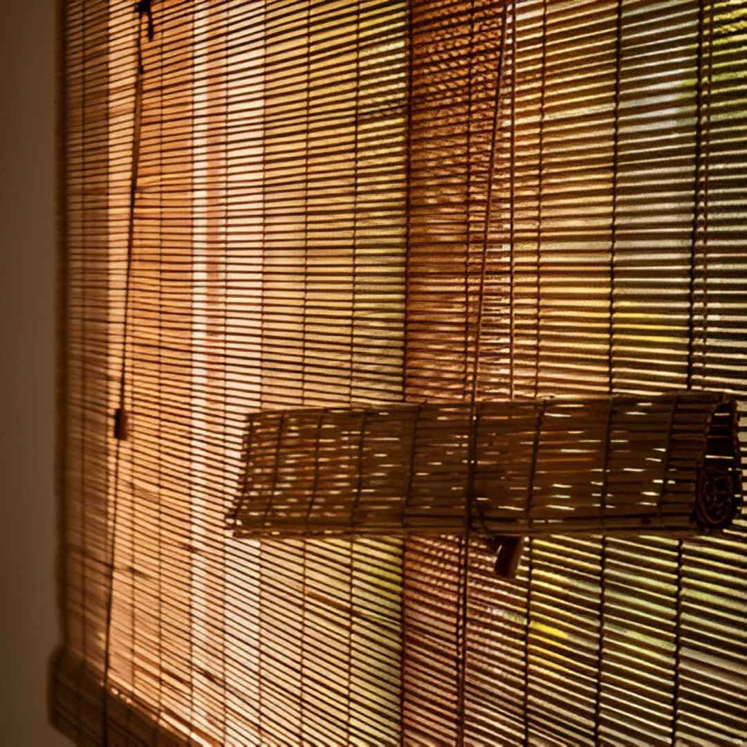 A close-up of a window covered with natural bamboo blinds. The thin bamboo slats have a woven texture, and warm sunlight filters through, creating a cosy and organic feel.