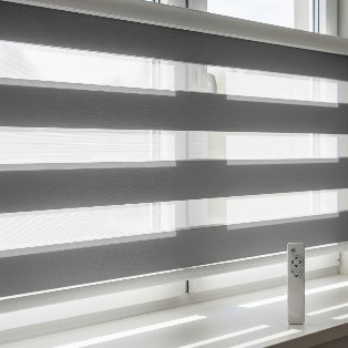 : A close-up view of motorised zebra blinds on a window. A remote control is visible on the windowsill, and the blinds' charcoal grey and white stripes are set to a position that allows light to filter in.