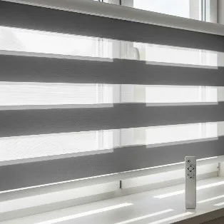 : A close-up view of motorised zebra blinds on a window. A remote control is visible on the windowsill, and the blinds' charcoal grey and white stripes are set to a position that allows light to filter in.